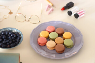 Plate of pastel macarons, cookies and chocolate, cup of tea of coffee, glass of bubble water, various berries, books and accessories on the table. Selective focus, pastel colors.