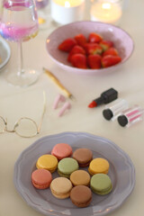 Plate of pastel macarons, cookies and chocolate, cup of tea of coffee, glass of bubble water, various berries, books and accessories on the table. Selective focus, pastel colors.