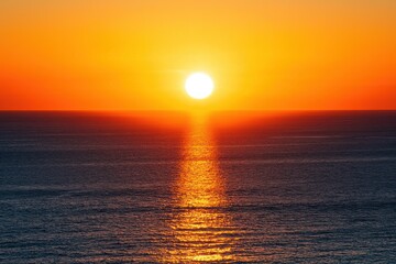 A serene sunset over the ocean with vibrant orange and red hues reflecting on the water at the beach, waves gently touching the shore under a sky filled with soft clouds