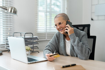 Senior Woman Shopping Online with Credit Card and Laptop While Talking on Phone at Home Office