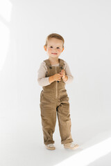 Child happy smiling boy in a pair of brown overalls stands in front of a white background