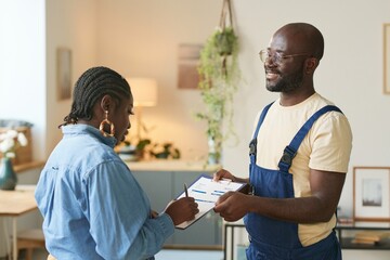 Obraz premium Side view portrait of smiling Black man in coveralls signing contract with female client holding clipboard in home interior