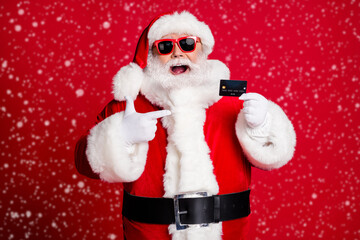 Photo of retired old man grey beard hold direct finger credit card open mouth smile offer good variant wear x-mas santa costume gloves coat belt sunglass cap isolated red color background