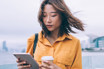 Beautiful Japanese hipster student checking received notification on modern touch pad standing at...