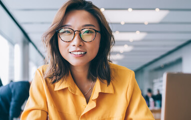 Portrait of Chinese positive hipster girl in trendy spectacles holding cellphone gadget for...