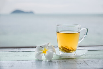 Cup of green tea with ginger servered with plumeria frangipani flower front of at sunset light on wooden tabletop. Sea view.