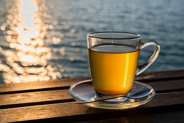 Cup of amber green tea  on wooden tabletop shining in the sunset light. Sea view.