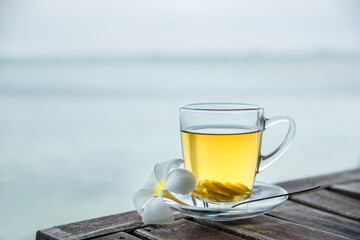 Cup of green tea with ginger servered with plumeria frangipani flower front of at sunset light on wooden tabletop. Sea view.