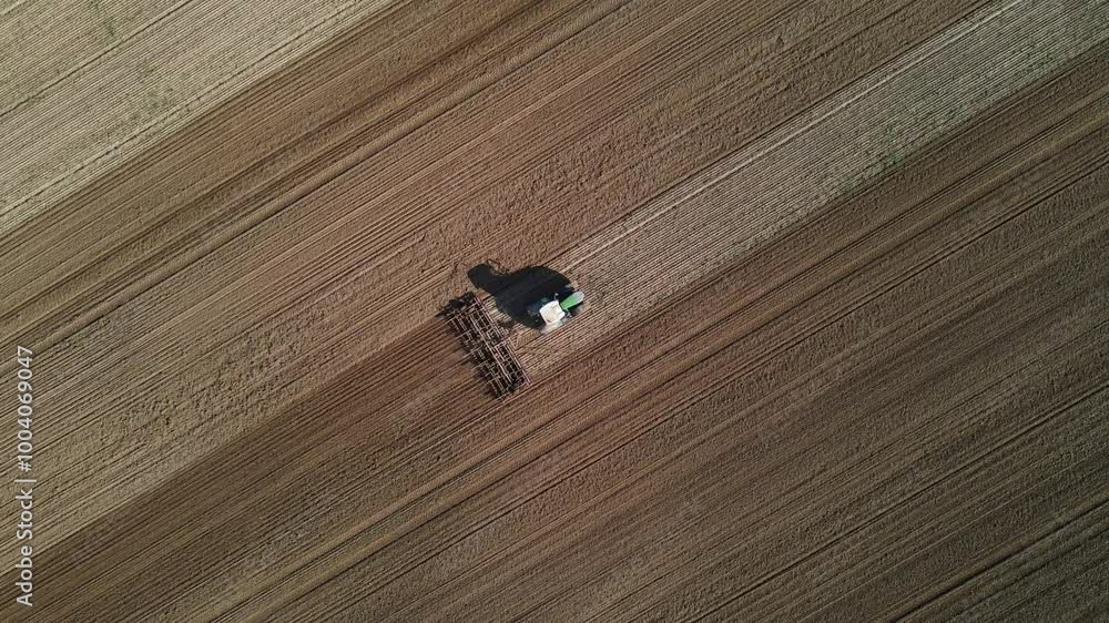 Poster an aerial view of a tractor working on a large agricultural field, plowing the soil in straight, par