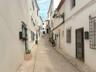 Fototapeta premium A narrow street in a European town with white buildings, windows with grills, lanterns, and a scooter.