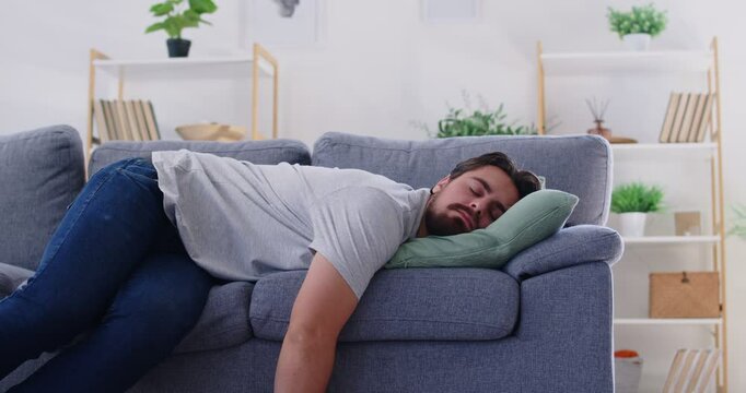 Tired young upset man lying on sofa in living room at home and sleeping after hard working day. Male person relaxing alone and having a rest on couch indoors. 4k video. Slow motion video.
