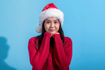 Asian woman dressed in a red knit sweater and wearing a Santa hat smiles sweetly while resting her fists against her cheeks. The background is a solid light blue, creating a cheerful holiday scene