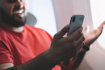 Selective focus on cellular which young smiling man holding in hands. Happy male flight passenger connecting to wireless internet on board sitting next to aircraft cabin window. Airplane Offline mode