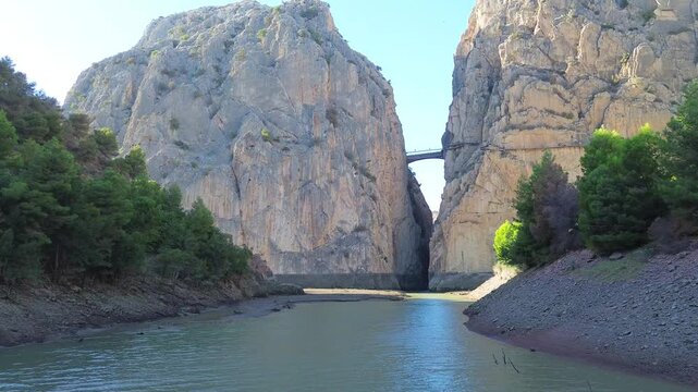 Drone flight over the river to the gorge Garganta del Chorro and Caminito del Rey, Spain. A bridge in a narrow gorge between huge high mountains, green forest on the river banks. 