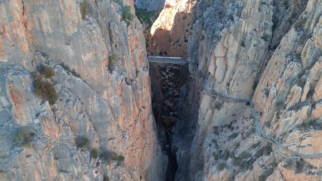 Aerial view from drone to the gorge Garganta del Chorro and Caminito del Rey, Spain. A bridge in a narrow gorge between huge high mountains and a river flows below. People are walking on the bridge.