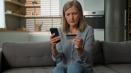 Angry confused senior Caucasian woman mature elderly female mother hold credit card security problem using bank app for payment retired grandparent mobile phone scam money loss failure error home sofa