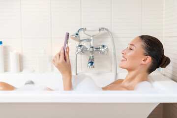 Happy young European lady relaxing in foamy bath with smartphone, reading beauty blog or scrolling social media feed and smiling, lying in bathtub, side view