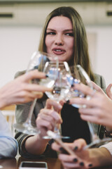 Young smiling woman saying toast during drinking white wine with female best friends on leisure time, happy cheerful hipster girls cheering with glasses and feeling good while sitting in cafeteria
