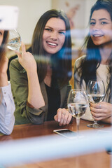Young smiling hipster girl holding modern smartphone gadget in hand and clicking selfie photo for posting at own web blog during leisure time with female friends for drinking wine and communicate