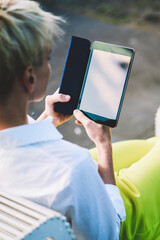 Back view of woman reading literature stories via modern e-book using 4g connection during rest on publicity area, hipster girl enjoying favourite bestseller via gadget in free time sitting outdoors