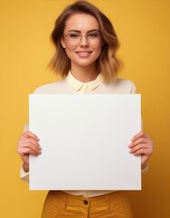 Fototapeta premium Smiling Young Woman with Glasses Holding a Blank White Poster for Advertisement on a Bright Yellow Background 