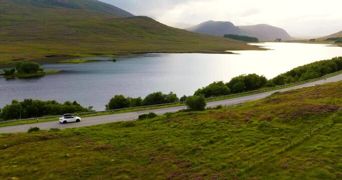 Aerial shot of white electro car driving with breathtaking Scottish landscape