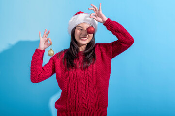 Obraz premium Asian woman dressed in a red knitted sweater and Santa hat holds gold and red Christmas ornaments near her ears. She stands against a light blue background, celebrating the festive holiday season