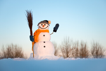 Funny snowman with broom and yellow scarf on snowy field. Blue sky on background