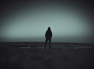 A person in hoodie in silhouette standing by the sea watching it.