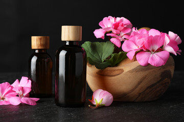 Bottles of geranium essential oil and beautiful flowers on black table, closeup
