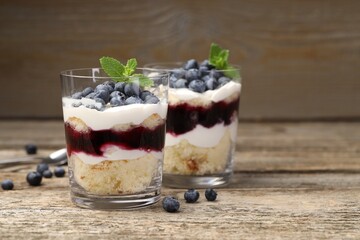 Tasty trifle dessert. Sponge cake, blueberries, jam and whipped cream in glasses on wooden table, closeup