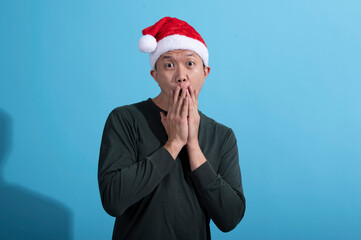 An Asian man wearing a red Santa hat and a dark green long-sleeved shirt expresses surprise, covering his mouth with both hands. He stands against a light blue background