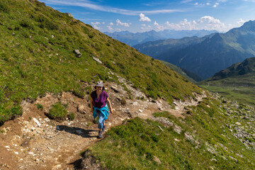 Fototapeta premium Woman on the way through beautiful mountain meadows with alpine autumnal flowers. the hiker on the footpath climbs the stony path up to the summit. beautiful alpine landscape with view over Austria