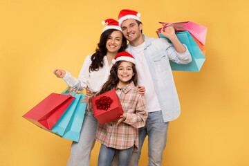 Xmas shopping. Portrait of European family in Santa hats buying presents, holding shopper bags on yellow studio background