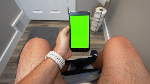 A man uses his touchscreen cell phone while sitting on a toilet. Green screen on unidentifiable device for custom content.	