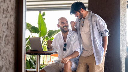 Two middle-aged male professionals are happily working on their laptops in the garden of a café on a summer day. Upon receiving some good news, they celebrate with joyful gestures and expressions