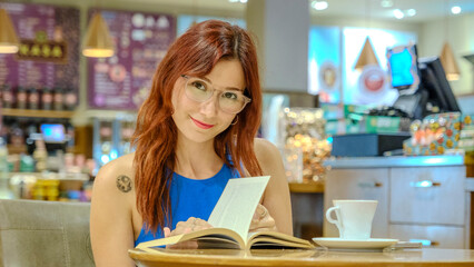 A beautiful young woman with red hair and glasses, wearing a blue t-shirt, is sitting in a lovely café on a summer evening, enjoying reading a book