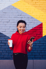 Cheerful Chinese female teenager with coffee beverage reading positive news in social networks connected to public 4g internet on mobile phone, happy Asian woman enjoying blogging, copy space area