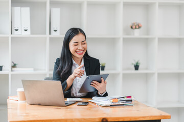 young woman in black suit smiles while using tablet at her desk, surrounded by office supplies and laptop. Her cheerful demeanor reflects productive work environment.