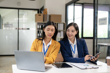 Two Asian women work online with laptop and smartphone. A joyful moment captured as two female colleagues laugh together during a productive meeting in the office.