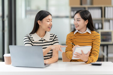 Obraz premium Two Asian women work online with laptop and smartphone. A joyful moment captured as two female colleagues laugh together during a productive meeting in the office.