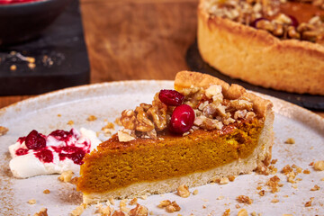 A slice of pumpkin pie with cream and cranberries rests on a ceramic plate on a wooden table