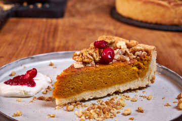 A slice of pumpkin pie with cream and cranberries rests on a ceramic plate on a wooden table