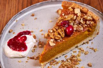 A slice of pumpkin pie with cream and cranberries rests on a ceramic plate on a wooden table