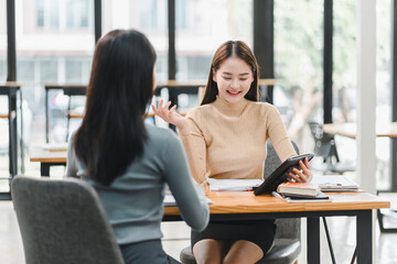 professional meeting takes place in modern office setting, featuring two women engaged in discussion. One woman is smiling while holding tablet, conveying positive and collaborative atmosphere.