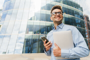 Business Professional Smiles While Holding a Laptop and Smartphone Outside an Office Building on a...