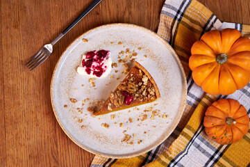 A slice of pumpkin pie on a ceramic plate rests on a wooden table with fall decor against a plaid towel background