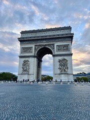 The Arc de Triomphe in Paris during the day with a dramatic, partly cloudy sky.  No cars, unrecognisable people in the background. 