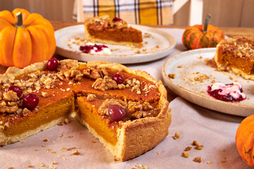 A slice of pumpkin pie on a ceramic plate rests on a wooden table with fall decor against a plaid towel background