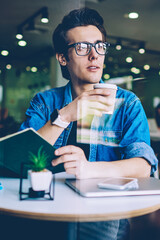 Pondering young man in eyeglasses enjoying caffeine beverage during learning indoors using knowledge literature for preparing to exam, thoughtful hipster guy casual dressed holding takeaway coffee